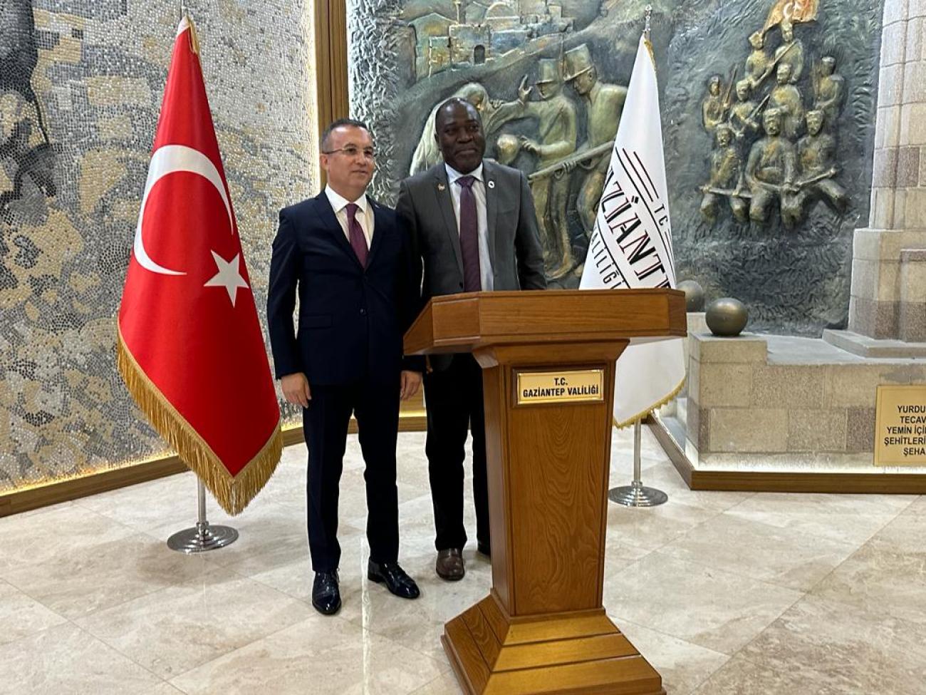 Governor of Gaziantep and RC Babatunde pose infront of a wall decoration. There is one Turkish flag and one flag which says Gaziantep behind them.